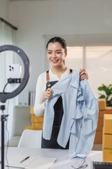 Young asian woman engaging in live stream commerce, presenting fashion apparel using a smartphone and ring light