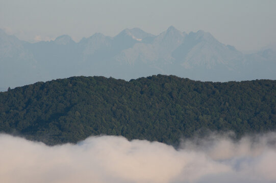 Aerial view of a sea of clouds rolling gently below a densely forested ridge, with distant, hazy mountain peaks piercing the skyline, Sivec, Kosice Region, Slovakia.