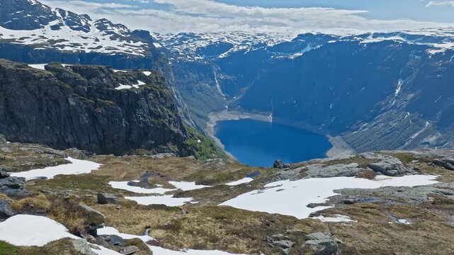 Snow-patched rocky plateau overlooking fjord at Trolltunga, Norway. Scenic mountain landscape with steep cliffs and calm blue water in bright summer light.