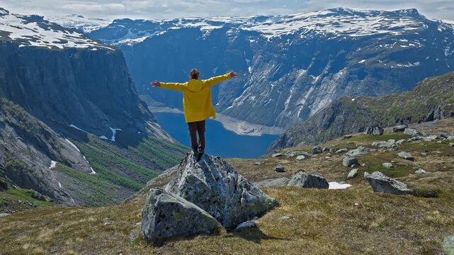 Woman Hiker standing on rock overlooking fjord at Trolltunga in Norway. Person in yellow jacket enjoying dramatic mountain cliffs and deep blue water during a summer hiking adventure.