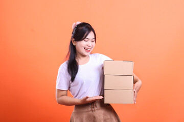 A cheerful woman holds a stack of cardboard boxes in her arms, ready for delivery, she has a big smile and a welcoming gesture, isolated on orange background