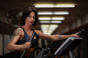 The athlete woman does exercises on an elliptical machine in a modern gym with dull lighting and focused on strength training