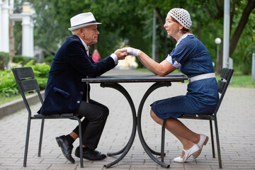Elderly couple engaging in a friendly arm wrestling match outdoors in a park setting with greenery and warm sunlight