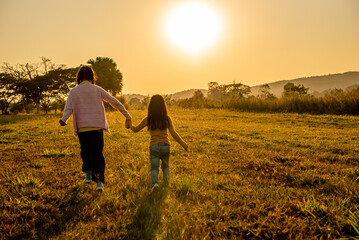 Fototapeta premium Rear view of two little sisters holding hands and walking through a grassy field at sunset, symbolizing friendship, childhood, and a bright future against a warm mountain backdrop.