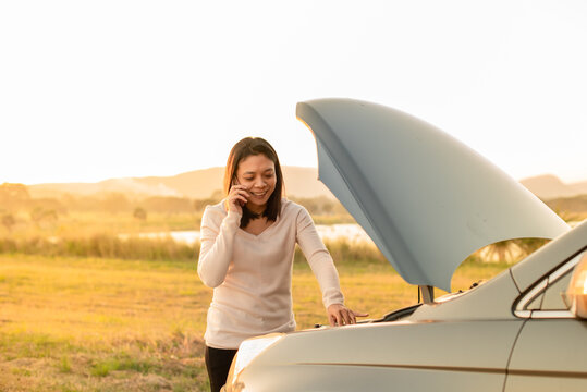 A woman smiling and looking relieved while talking on her mobile phone next to her car with the hood open, suggesting a successful call for help or fixing a minor engine problem.