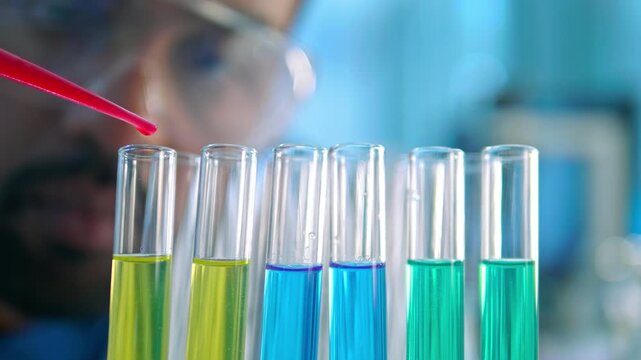 A scientist in a lab coat and safety goggles carefully dispenses a red liquid into a row of test tubes containing various colored solutions. The setting suggests a controlled laboratory environment.