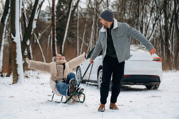 Joyful couple enjoying winter fun, with woman on sled being pulled by man in snowy forest, showcasing playful outdoor activities and seasonal happiness