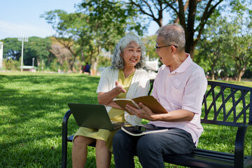 Happy senior Asian couple sitting on a park bench using a laptop and notebook while talking in a sunny green park.