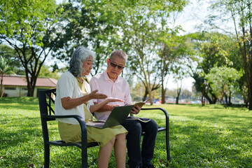 Elderly Asian couple using a laptop and reading a book while sitting on a park bench in a sunny green garden.