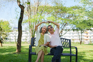 Happy senior couple sitting on a park bench and making a heart shape with their hands in a sunny public park.