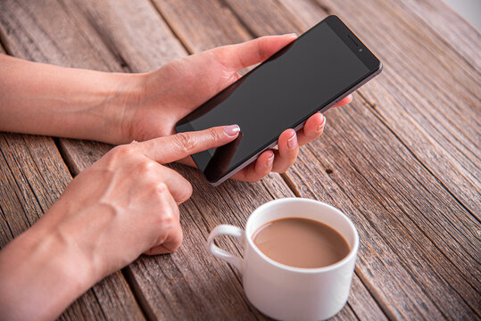 A person holding a black smartphone over a rustic wooden table next to a cup of coffee.