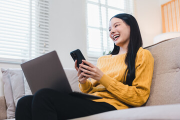 Young Asian woman laughing while using her smartphone and laptop sitting on a sofa in a bright living room