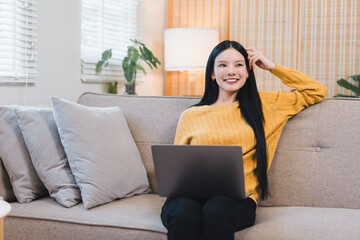 Happy Asian woman using a laptop and smiling while sitting on a sofa in a bright living room home office.