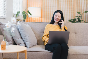 Smiling young Asian woman working from home using laptop and talking on phone while sitting on a sofa