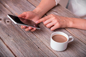 A woman uses a white smartphone while sitting at a rustic wooden table next to a cup of coffee.