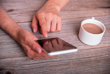 A woman uses a white smartphone while sitting at a rustic wooden table next to a cup of coffee.
