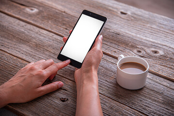 A woman holds a smartphone with a blank white screen next to a cup of coffee on a rustic wooden table.