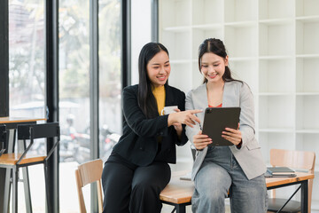 Two professional Asian business women collaborating and discussing work while looking at a digital tablet in a bright office.