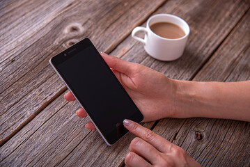 A person holding a black smartphone over a rustic wooden table next to a cup of coffee.