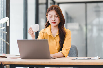 Professional young Asian businesswoman working on her laptop and holding a coffee cup in a bright modern office