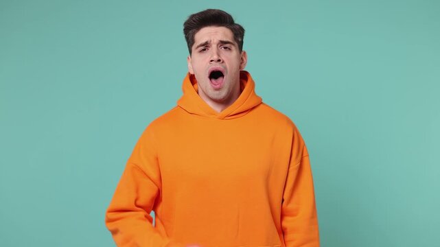 Tired sad young man he wears orange hoodie did not get enough sleep last night after party and barely got up in the morning yawn isolated on plain pastel light blue cyan background. Lifestyle concept