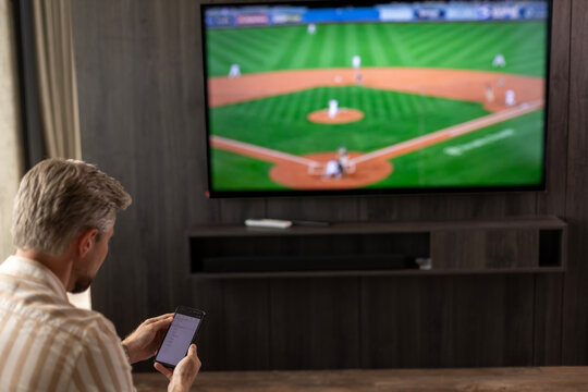 Caucasian man holding smartphone with sports bets app while watching baseball match at home in living room