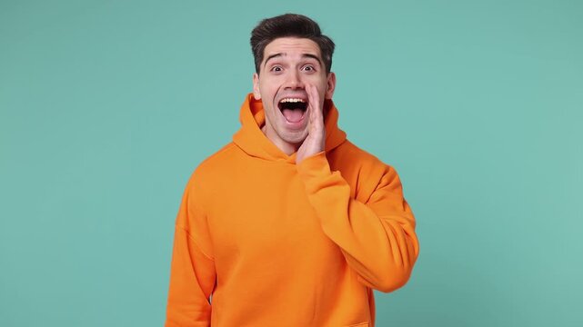 Excited happy fun young man he wears orange hoodie scream hot news about sales discount with hands near mouth hurry up isolated on plain pastel blue cyan background studio portrait. Lifestyle concept