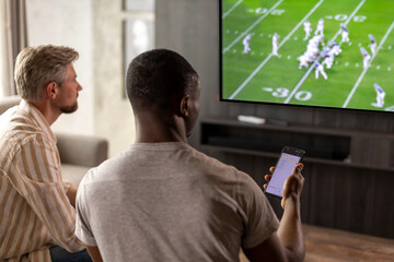 Two friends holding smartphone with sports bets app while watching American football match at home in living room.