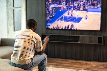 African man holding smartphone with sports bets app while watching basketball match at home in living room.