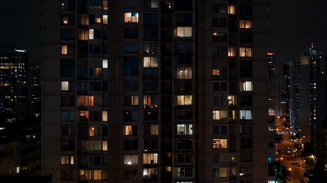 Night view of urban apartment building with lights on in different windows and city lights shining in the background during nighttime in a metropolitan area