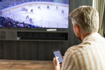 Caucasian man holding smartphone with sports bets app while watching ice hockey match at home in living room