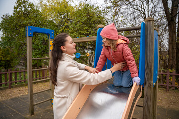 Obraz premium Mother Guiding Daughter Down Playground Slide Careful Hands On Jacket, Shiny Metal Slide, Autumn Leaves, Supportive Posture, Smiling Trustful Moment