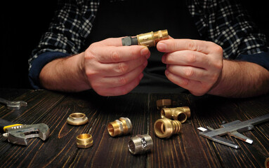 Two hands work with brass fittings on a wooden table. Various tools and fittings are scattered around. The activity focuses on plumbing repair. Evening light enhances the scene