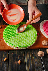 A person prepares a dessert by spreading condensed milk on a green cake. Ingredients such as nuts and red cake are placed nearby. The photo shows a workstation busy with food preparation.