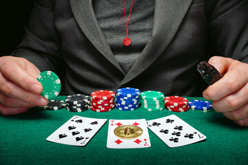 On a green table lie three playing cards with bitcoin and some chips. A man holds a red chip, ready to place a bet during a late-night poker game.