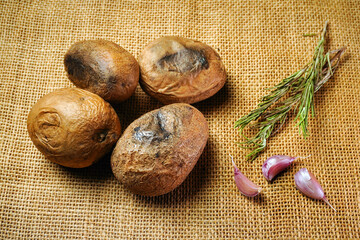 Four brown baked potatoes and several garlic cloves sit on a burlap cloth with herbs arranged nearby, ready for meal preparation in a kitchen setting