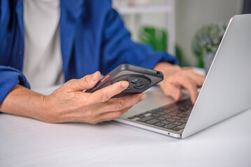 Close up of senior woman hands holding smartphone and typing on laptop keyboard. Elderly businesswoman multitasking with technology, data entry, and digital connection concept.