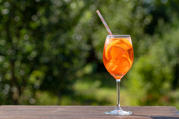 Aperol spritz cocktail in wine glass with drinking straw on nature background. Classic italian aperitif aperol spritz cocktail with orange and ice cubes. Summer refreshing drink with sunlight