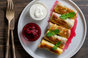 Fried rolled pancakes stuffed with cottage cheese and cherry, sweet dessert on plate, closeup, top view. Homemade baking. Delicious fast food for breakfast. Wrapped pancakes with sour cream and jam