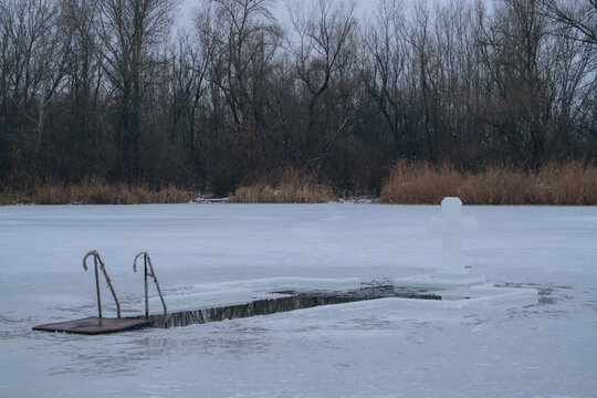 Symmetrical view of a cross-shaped ice hole and a large ice cross standing on a frozen river. Wintry landscape with bare trees and a gray overcast sky in the background. Concept of the Orthodox Epipha