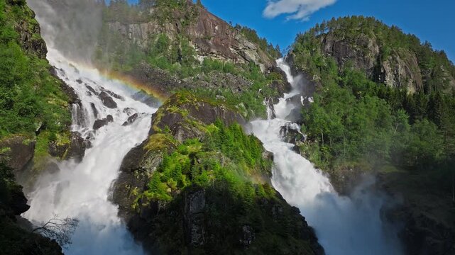 Latefossen waterfall with rainbow mist in Norway. Aerial view of powerful twin cascades surrounded by lush green forest on a sunny summer day.
