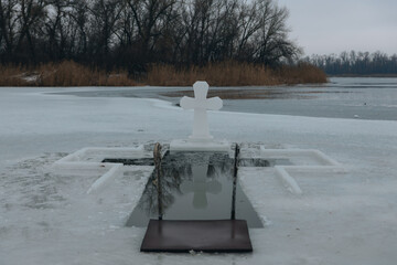 Symmetrical view of a cross-shaped ice hole and a large ice cross standing on a frozen river. Wintry landscape with bare trees and a gray overcast sky in the background. Concept of the Orthodox Epipha