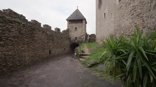 Stone walkway leads to a defensive tower with a pointed roof inside a medieval castle in Italy. Historic stone walls and green plants under an overcast sky create a moody atmosphere.