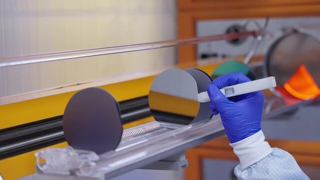 Gloved technician inspecting reflective silicon wafers during semiconductor fabrication, with thin film coatings and precision handling inside a cleanroom process station