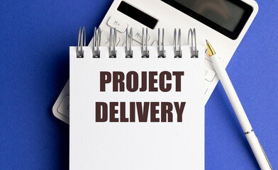 Project delivery written on spiral notepad with pen and calculator on blue background