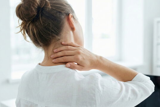 Woman suffering from neck pain and shoulder tension while working in a bright office, massaging her painful neck for relief.