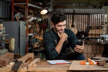 Smiling luthier reading message on smartphone in workshop, small business craftsman at workbench