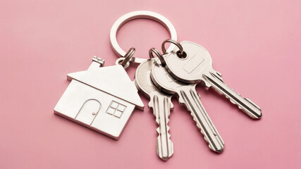 Keys are placed on a pink background with a silver keychain featuring a house design, symbolizing home ownership and the start of new chapters in life