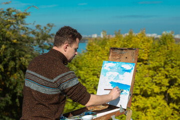 An artist painting a landscape on a canvas set on an easel outdoors, surrounded by nature.