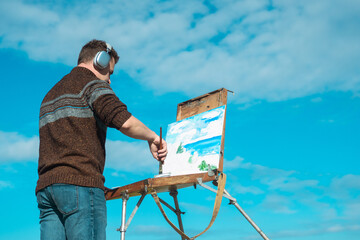 An artist in headphones painting a landscape on a canvas set on an easel outdoors, surrounded by nature.